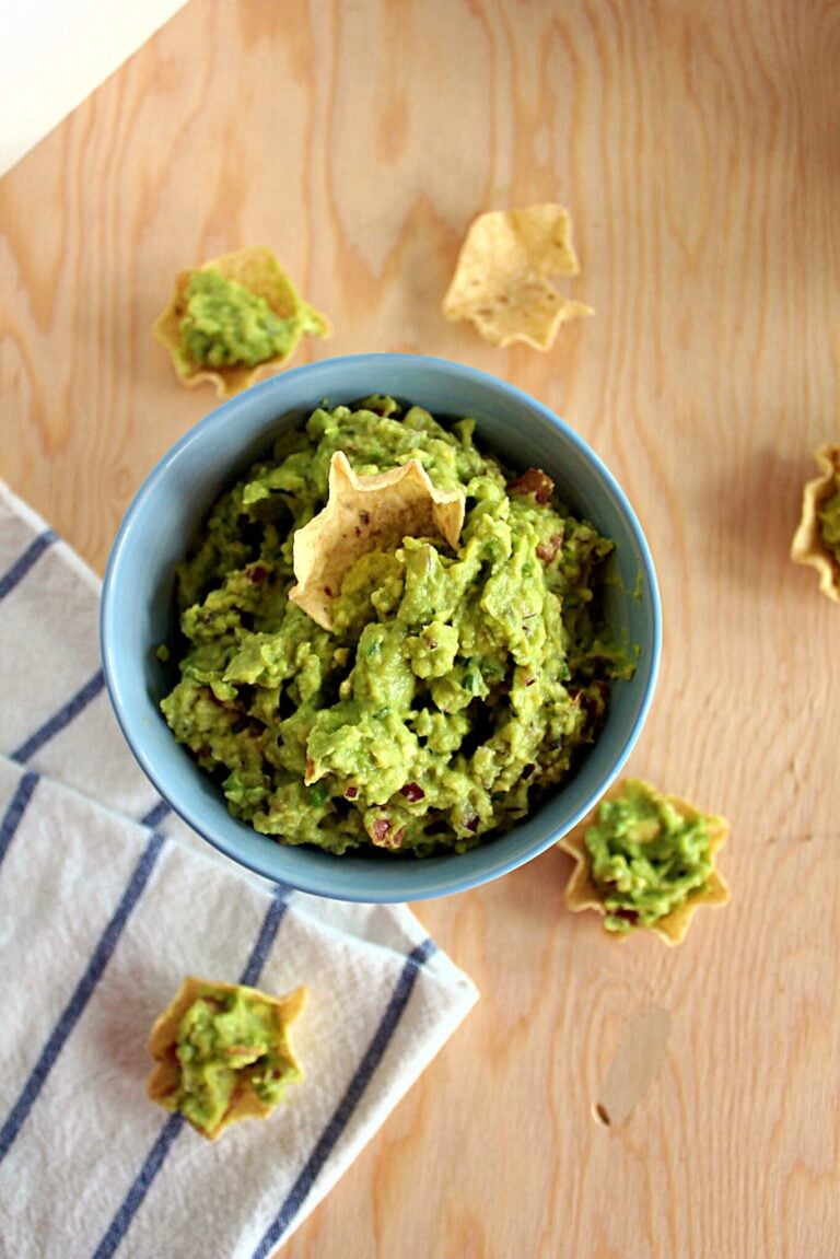 a beige tortilla chip scooping up bright green guacamole dip in a light blue bowl against a wood background