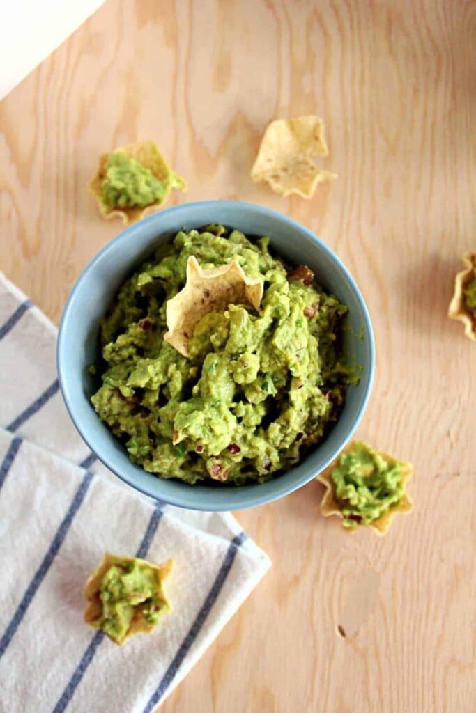 a beige tortilla chip scooping up bright green loaded guacamole dip in a light blue bowl against a wood background