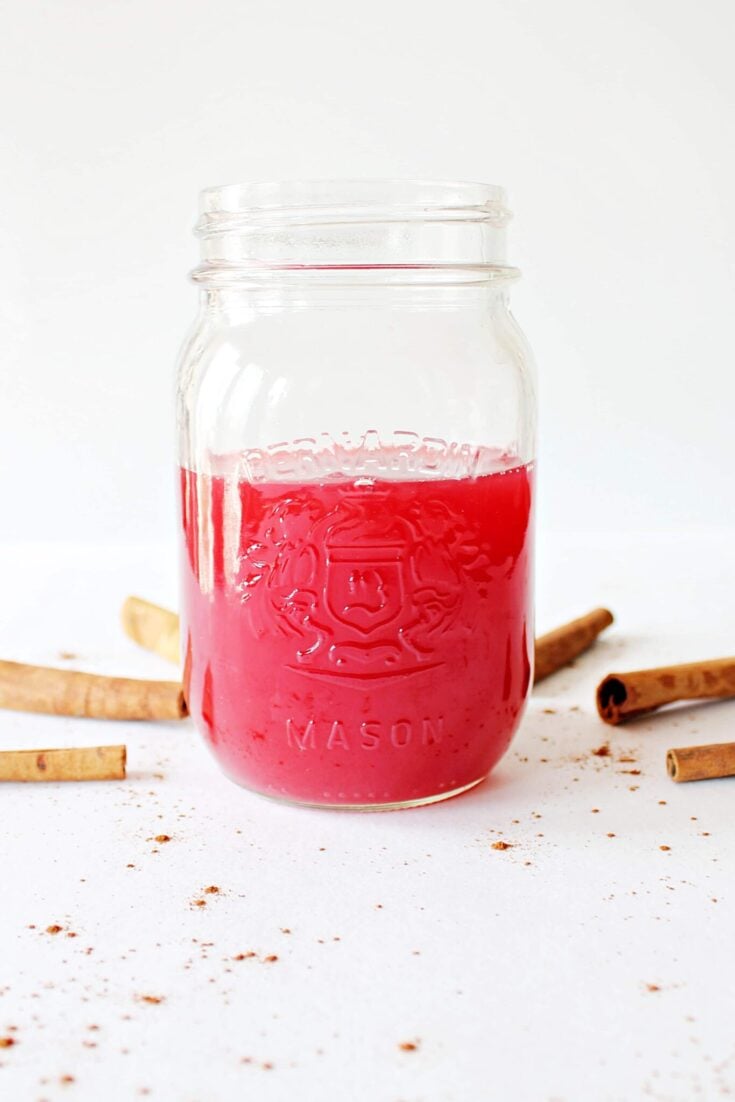 A jar half filled with bright pink rhubarb syrup. There are brown cinnamon sticks arranged around the jar.