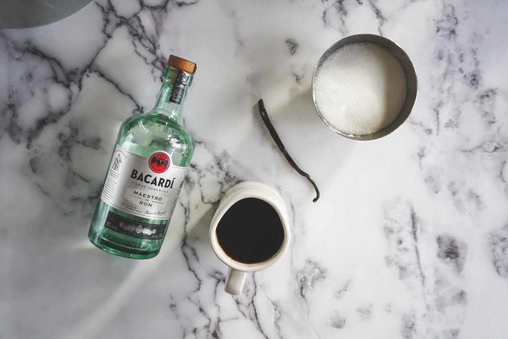 Looking down at all the ingredients needed for sous vide kahlua. A bottle of white Bacardi rum, a mug of coffee, a bowl of sugar and a vanilla bean.