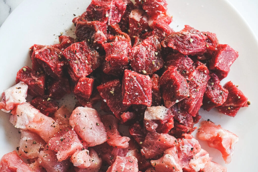 A close up of the cubed beef and pork before being browned. The meat is dotted with coarse ground salt and pepper.