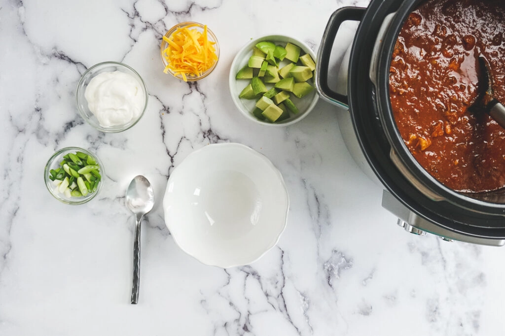 Cooked keto instant pot chili in the Instant Pot being ladled into a white bowl with toppings spread around the bowl.
