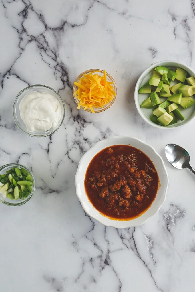 Dark red keto chili in a white bowl surrounded by the different topping suggestions; diced avocado, shredded cheese, sour cream, and sliced green onion