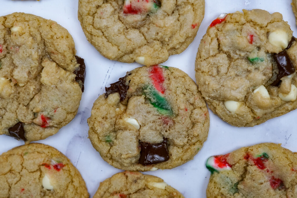 Candy cane cookies spread on the counter. Can see chunks of candy cane, white chocolate chips, chunks of chocolate.