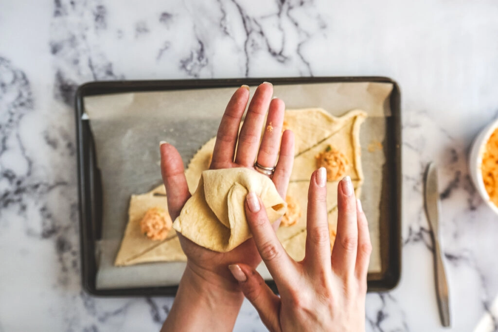 Hands folding in the edges of a triangle crescent roll around the buffalo chicken filling.