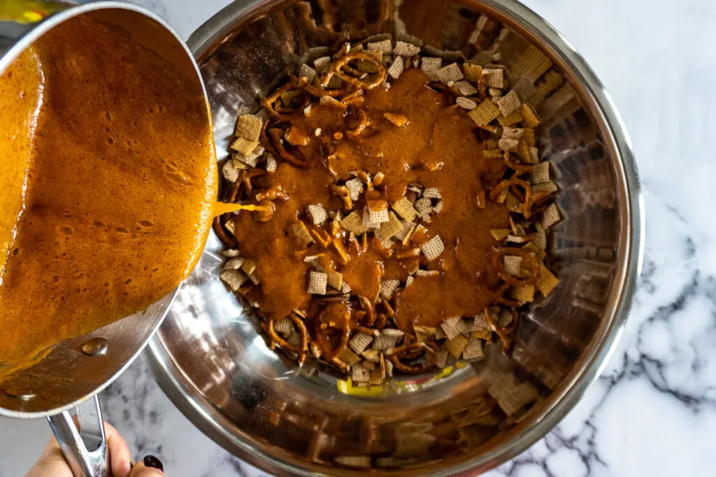 Pouring sriracha caramel on the chex, life, pretzels, and nuts in a large stainless steel bowl.