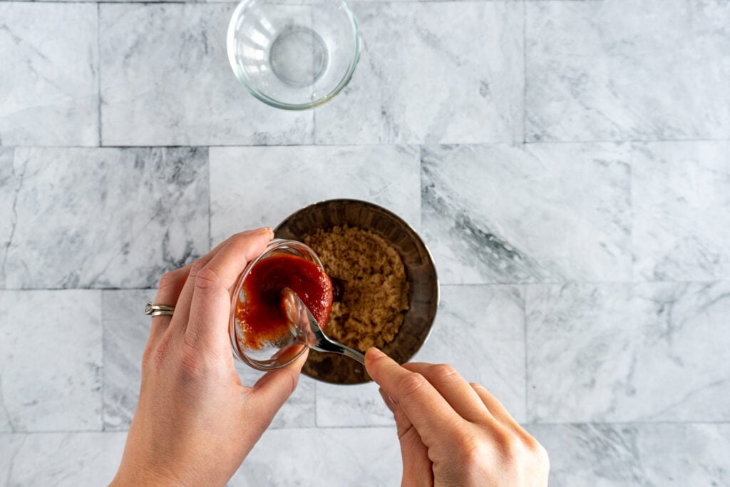 Pouring sriracha from glass bowl into bowl with brown sugar..