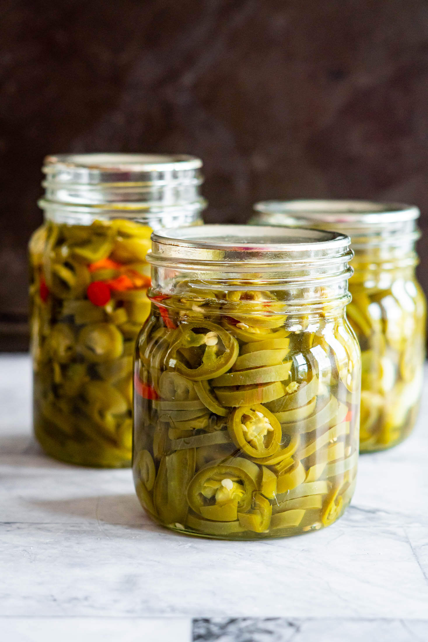 Three pint sized jars of water bath canned pickled jalapeños on a marble countertop. 